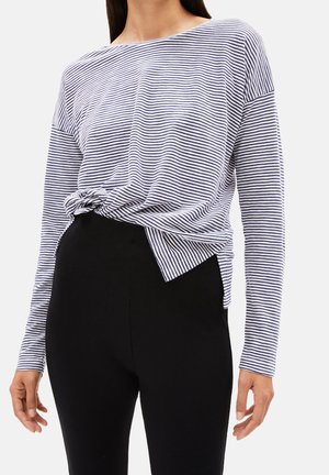 Kobieta w granatowo-białej bluzce w paski z długim rękawem, zawiązanej w pasie, w połączeniu z wysokim stanem czarnych spodni, na jednolitym tle.