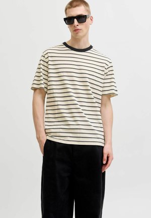 Jeune homme portant des lunettes de soleil noires, un t-shirt à manches courtes beige et noir à rayures horizontales et un pantalon noir ample, les mains dans les poches.