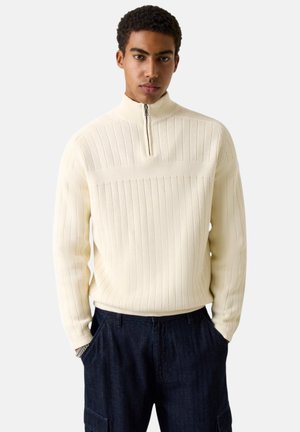 Jonge man die een roomkleurige geribbelde gebreide trui draagt met een kraag met rits en donkerblauwe broek, staand met handen in de zakken.