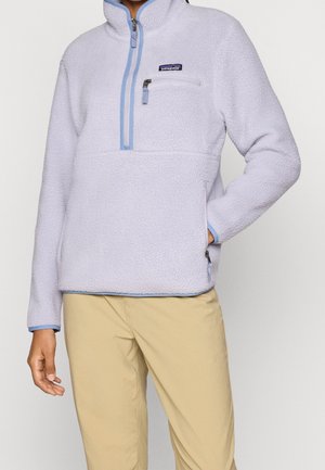 Osoba nosząca lawendowy polar z zamkiem do ćwiartki i kieszenią na piersi, połączony z beżowymi spodniami, ręka w bocznej kieszeni.