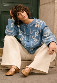 Ženska kodrastih las v modri vezeni jakni, kremnih širokih hlačah in rjavih čevljih z visokim petom sedi na stopnici ob zelenih vratih in kamnitem zidu.