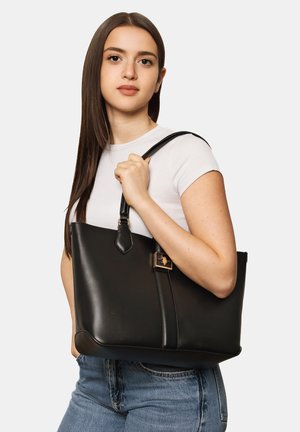 Jeune femme aux longs cheveux bruns, en t-shirt blanc et jean, porte un grand sac cabas en cuir noir sur l’épaule.