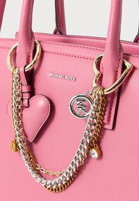 Ροζ δερμάτινη τσάντα διαθέτει χρυσά μεταλλικά στοιχεία, ασημένιες και χρυσές αλυσίδες, μπρελόκ σε σχήμα καρδιάς και λογότυπο στο μπροστινό μέρος.
