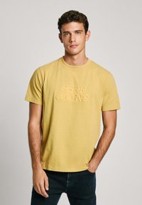 Geltona medvilninė marškinė su trumpomis rankovėmis, centre, su tekstūruotu logotipu "PEPE JEANS". Klasikinio apykaklės dizaino.
