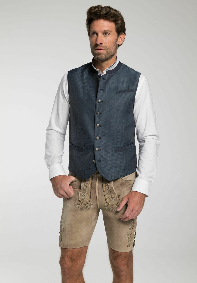 Blau gemusterte Weste mit hohem Kragen und silbernen Knöpfen, kombiniert mit hellbraunen Shorts mit bestickten Details und Knopfakzenten.