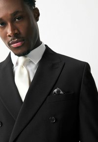 Chaqueta de traje negra de doble botonadura con dos botones, complementada con una camisa blanca y una corbata marfil. Presenta un pañuelo de bolsillo negro estampado.