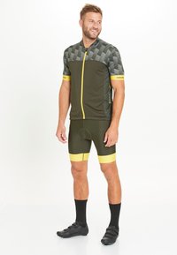 Maillot de cyclisme vert avec un haut à motifs et des accents jaunes, associé à un short vert assorti avec des rayures jaunes. Chaussures de cyclisme noires.