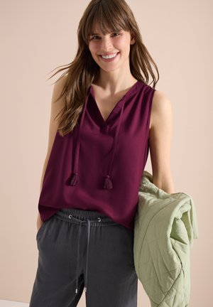 Femme souriante portant un haut bordeaux sans manches à franges et un pantalon foncé à cordon, tenant une veste matelassée vert clair devant un fond uni.