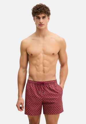 Modèle masculin aux cheveux bouclés portant un short de bain rouge à motifs, debout avec une main dans la poche sur un fond clair uni.