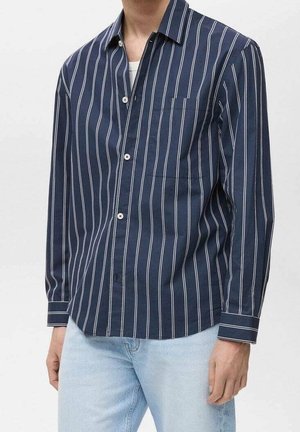 Mężczyzna ubrany w granatową koszulę zapinaną na guziki z białymi pionowymi paskami oraz jasnoniebieskie dżinsy, stojący na tle jednolitego tła.