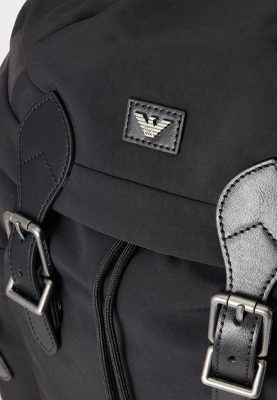 Sac à dos noir avec des boucles argentées, des sangles en cuir noir et un patch logo aigle Armani en métal sur le rabat en tissu.