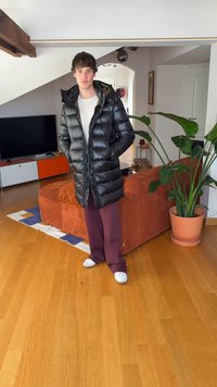 Zwarte pufferjas met een glanzende afwerking, lange mouwen, geribbelde accenten, gedragen over een licht shirt, gecombineerd met bordeauxrode broek en witte schoenen.