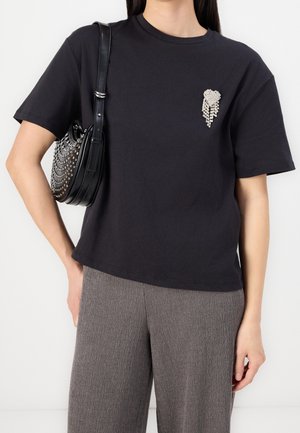 Kobieta ubrana w czarny t-shirt z srebrną ozdobną broszką, szare teksturowane spodnie i czarną ćwiekowaną torbę na ramię.