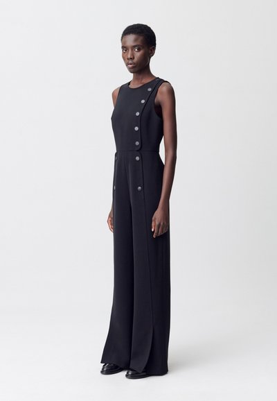 Combinaison noire sans manches, avec des boutons sur le devant et des jambes larges. Tissu lisse avec un léger éclat et une coupe structurée. Chaussures noires.