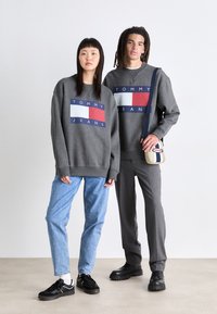 Sivé mikiny s červeným a modrým blokovým dizajnom s označením "TOMMY JEANS," v kombinácii s modrými džínsami a tmavou obuvou, na neutrálnom pozadí.