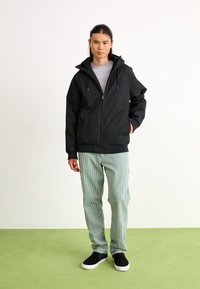 Black, zippered jacket with a hood, featuring elastic cuffs and a loose fit. Worn over a gray shirt and paired with striped green pants and black sneakers.