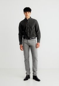 Νέος άνδρας που στέκεται, φοράει μαύρο πουκάμισο με κουμπιά, ανοιχτό γκρι τζιν με ζώνη και μαύρα δερμάτινα μποτάκια αστραγάλου σε λευκό φόντο.