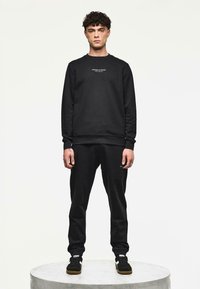 Uomo in piedi su una piattaforma rotonda che indossa un felpa nera con il testo "WEEKEND OFFENDER", pantaloni neri abbinati e sneakers nere con strisce bianche.