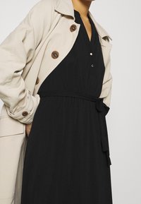 Frau, die einen beigen Trenchcoat über einem schwarzen, mit Gürtel versehenen Kleid mit Knöpfen trägt, die Hände in den Manteltaschen, vor einem schlichten Hintergrund.