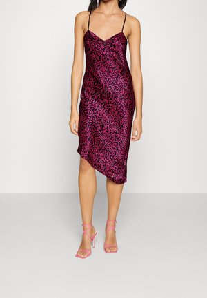 Femme portant une robe nuisette sans manches de couleur violet foncé avec des motifs abstraits roses et des sandales à talons hauts à brides roses, debout devant un fond uni.