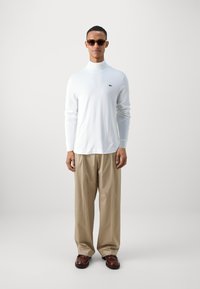 Chemise blanche à manches longues avec col roulé et logo vert, associée à un pantalon beige clair et des chaussures marron. Tissu lisse, coupe décontractée.