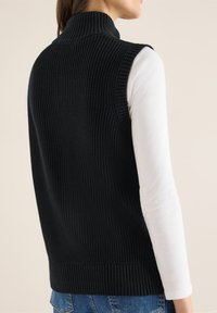 Černá žebrovaná pletená vesta s vysokým límcem, která má strukturovaný vzor a boční rozparky, nošená přes bílý dlouhý rukáv.