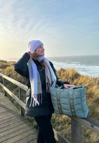 Zwarte pufferjack, lichtpaarse gebreide beanie en een grijze sjaal. Houdt een zachte, gevoerde mintgroene draagtas met logo en handvatten vast.