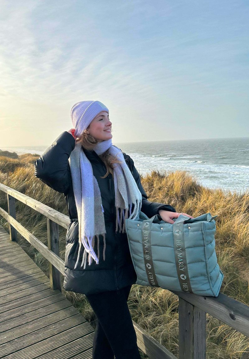 Zwarte pufferjack, lichtpaarse gebreide beanie en een grijze sjaal. Houdt een zachte, gevoerde mintgroene draagtas met logo en handvatten vast.