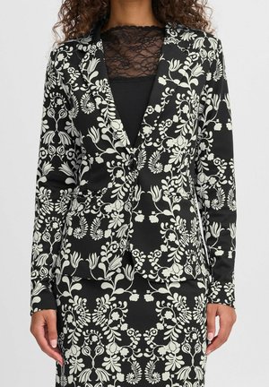 Kobieta ubrana w czarną marynarkę i spódnicę z białym wzorem kwiatowych pnączy oraz czarną koronkową bluzkę pod spodem.