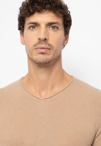 Taillierter beigefarbener V-Ausschnitt-Pullover mit gerippter Textur. Weicher Stoff, glatte Oberfläche und minimalistische Nähte. Sauberer Hintergrund, der das Kleidungsstück hervorhebt.