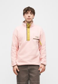 Νέος άνδρας που φοράει ροζ φλις πουλόβερ με κίτρινη λωρίδα κουμπιών και τσέπη στο στήθος με φερμουάρ, συνδυασμένο με σκούρο καφέ παντελόνια, στέκεται σε ουδέτερη στάση.