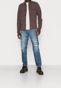 Hnědá flanelová košile se dvěma předními kapsami, pod ní béžové tričko, opotřebované modré džíny a černé šněrovací boty.