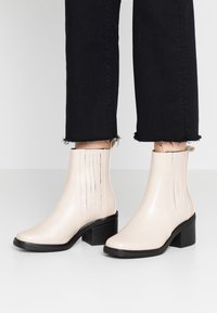 Bottes de cheville beiges avec une finition en cuir lisse, un talon bloc, des panneaux élastiques sur les côtés avec des coutures verticales et une semelle noire texturée.