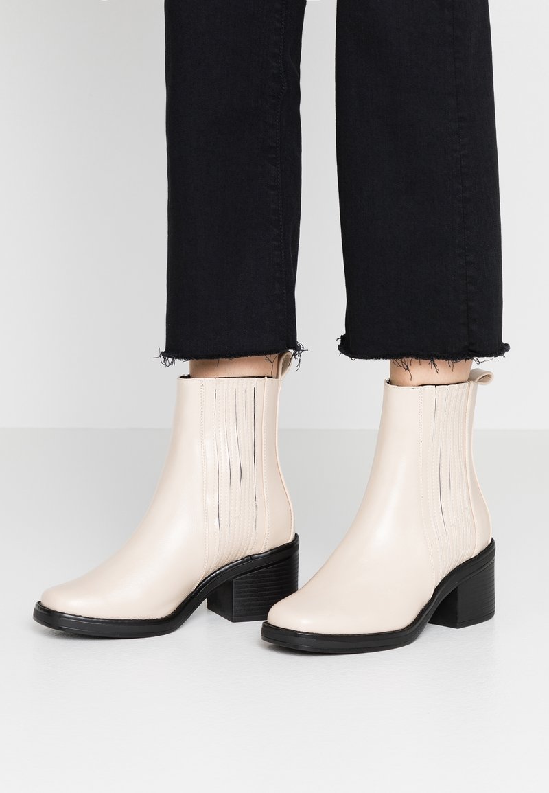 Bottes de cheville beiges avec une finition en cuir lisse, un talon bloc, des panneaux élastiques sur les côtés avec des coutures verticales et une semelle noire texturée.