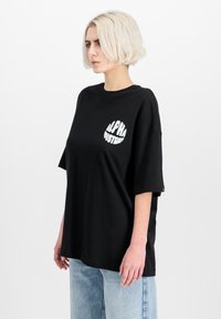 Černé oversized tričko s krátkými rukávy, s bílým logem "ALPHA INDUSTRIES" na hrudi, vyrobené z bavlněné tkaniny.