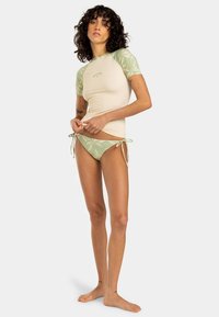 Világos bézs rövid ujjú fürdőfelső zöld levélmintával a vállakon, hozzáillő zöld oldalt megkötős bikini alsóval.