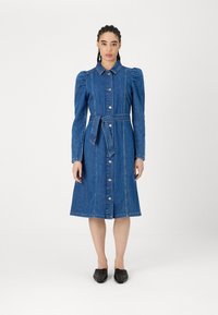 Vestido de ganga com frente abotoada, gola e cinto de laço. Apresenta mangas longas, ombros bufantes e um comprimento até ao joelho. Textura suave.