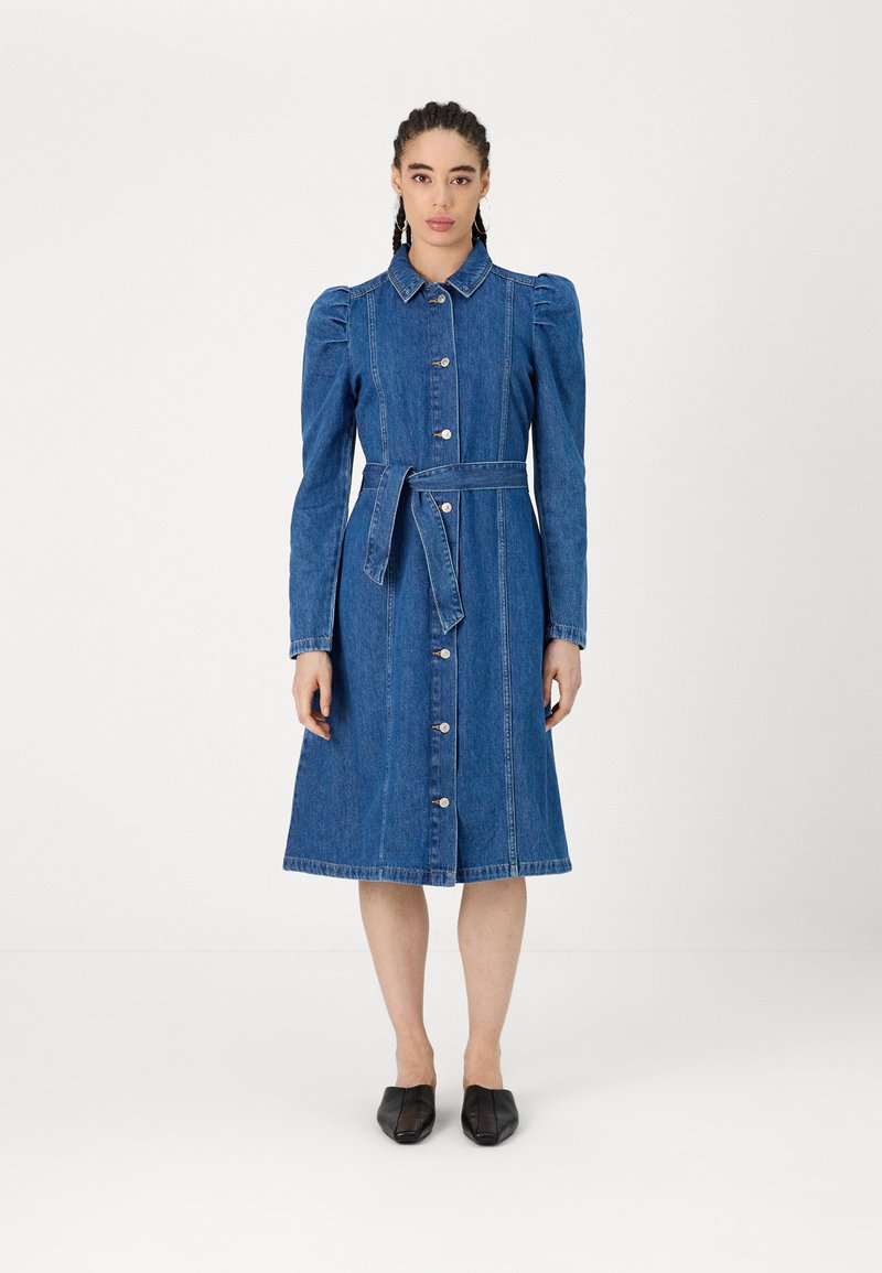 Vestido de ganga com frente abotoada, gola e cinto de laço. Apresenta mangas longas, ombros bufantes e um comprimento até ao joelho. Textura suave.