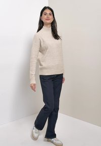 Femme aux cheveux foncés portant un pull beige à col roulé, un jean bleu foncé et des baskets blanches, debout avec une jambe croisée dans un coin du studio.