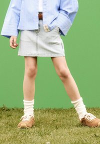 Mini-jupe en denim avec un délavage, associée à une chemise rayée bleu clair, des baskets marron et des chaussettes blanches tendance sur un fond d'herbe verte.