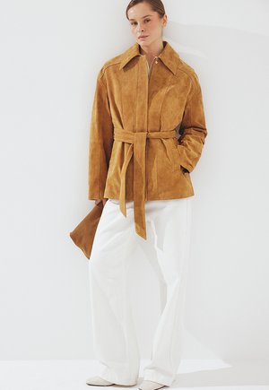 Ženska, oblečena v mentolno zunanjo jakno z ovratnikom, bele široke hlače, v roki drži ujemajočo torbico iz mentolnega semiša na belem ozadju.