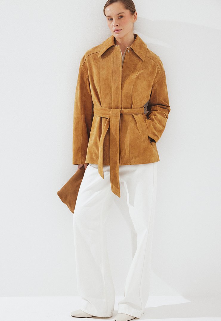 Žena oblečená v béžovom semišovom páse a biele široké nohavice, drží zladenú béžovú semišovú kabelku na bielom pozadí.