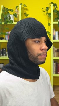Black ribbed knit hooded scarf with a snug fit. Features a long neck section. Background shows yellow walls and green plants.
