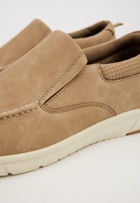 Zapatos slip-on beige hechos de cuero texturizado, con costuras decorativas y una suela de goma flexible con patrones de tracción.