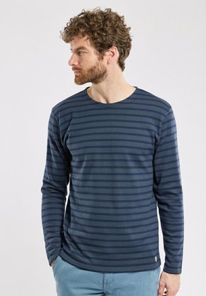 Navy long-sleeve shirt with horizontal light blue stripes, round neckline, and a small logo tag at the bottom left hem. Soft cotton blend fabric.