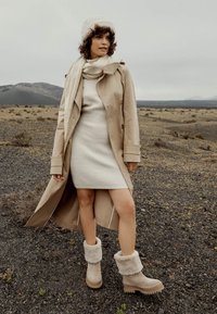 Abrigo de trench beige, vestido de punto color crema y botas beiges forradas de piel con puños texturizados en un terreno rocoso bajo un cielo nublado.