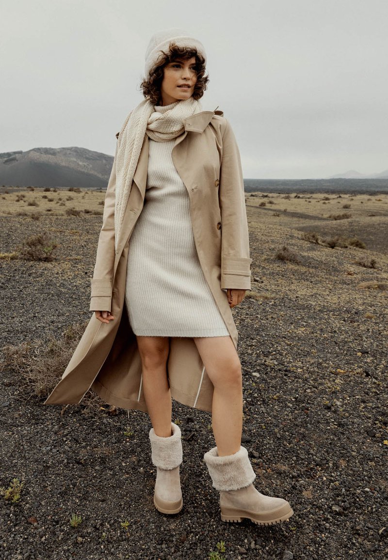 Abrigo de trench beige, vestido de punto color crema y botas beiges forradas de piel con puños texturizados en un terreno rocoso bajo un cielo nublado.