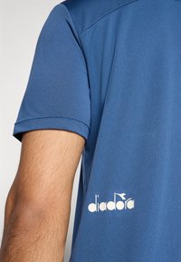 Blaues Sportshirt aus atmungsaktivem Material, mit kurzen Ärmeln, Rundhalsausschnitt und einem weißen "diadora"-Logo auf der linken Seite.