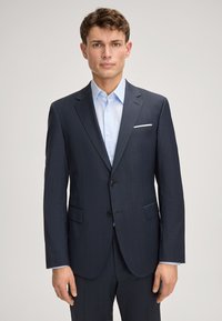 Granatowy garnitur dopasowany, z jednorzędową marynarką, klapami w kształcie notcha, dwoma przednimi kieszeniami i jasnoniebieską koszulą pod spodem.