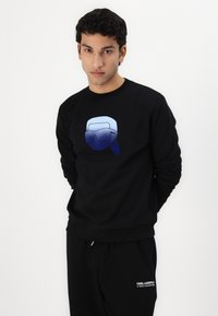Black sweatshirt with a gradient blue graphic of a figure wearing glasses. Features a round neckline and cuffed sleeves. Paired with black joggers.